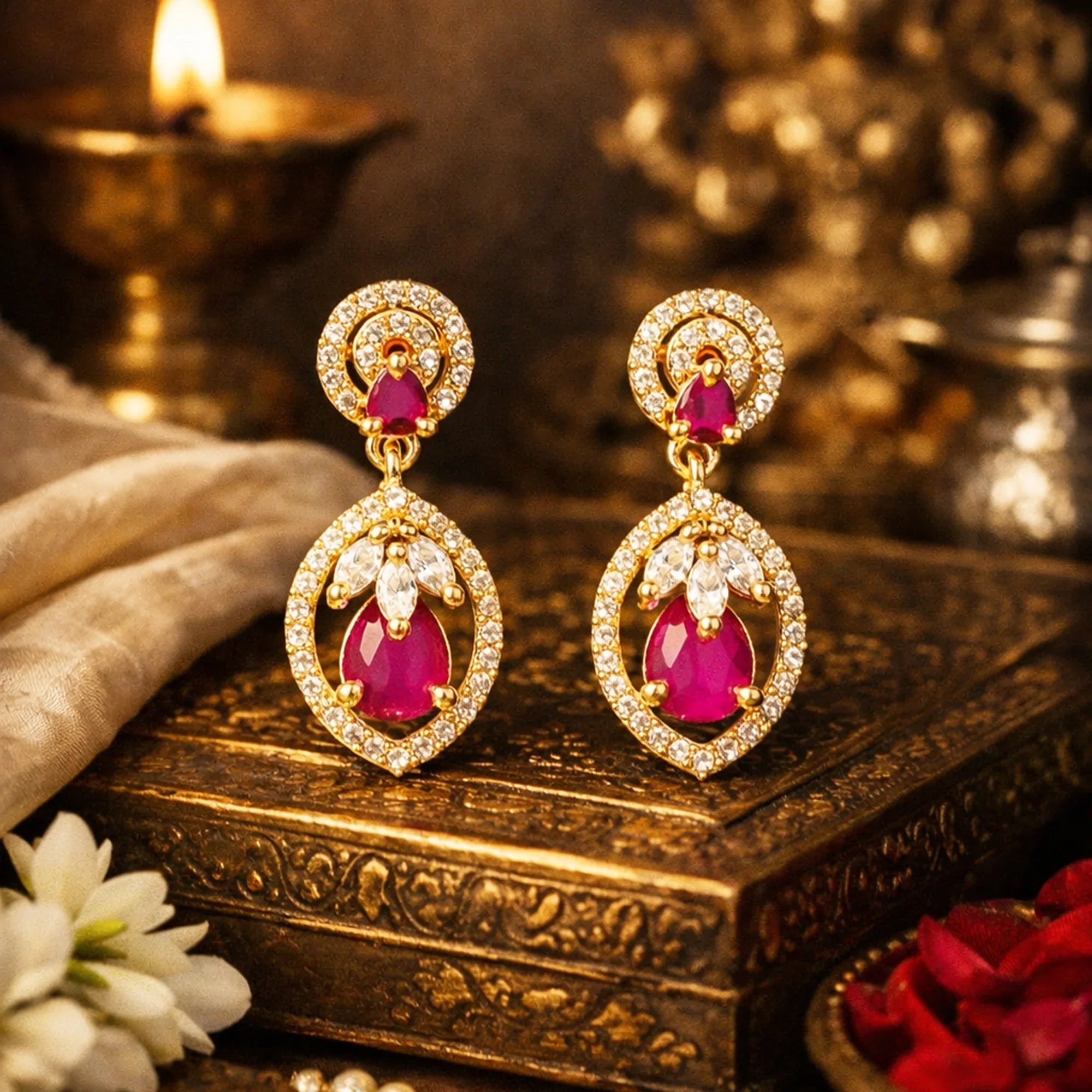 Gold earrings with red gemstones on a decorative box with flowers and candles in the background