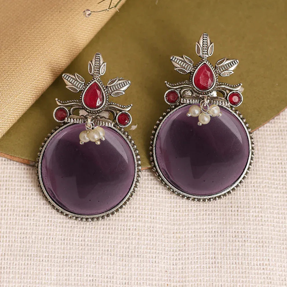 Purple earrings with red gemstones and silver accents on a textured surface