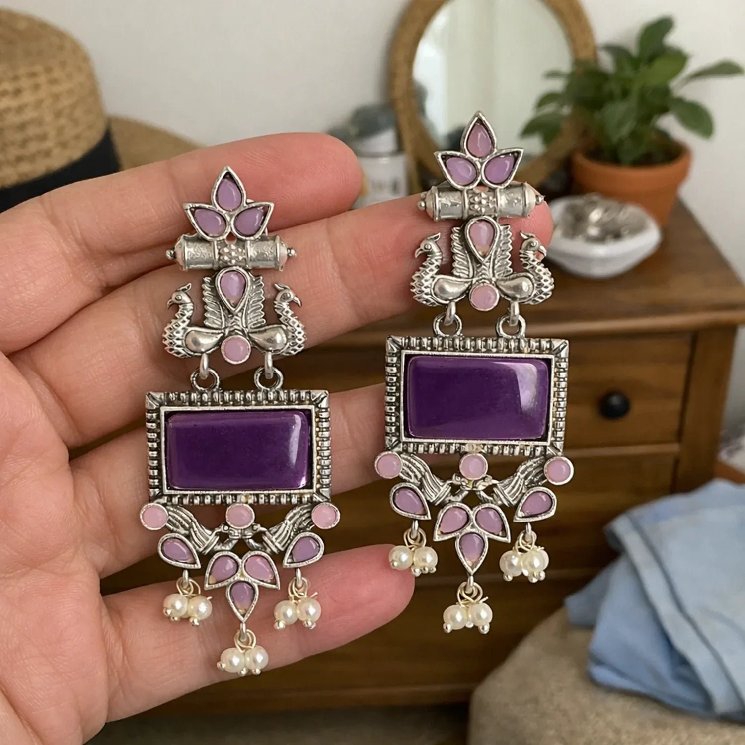 Ornate earrings with purple stones held in a hand, with a blurred indoor background.