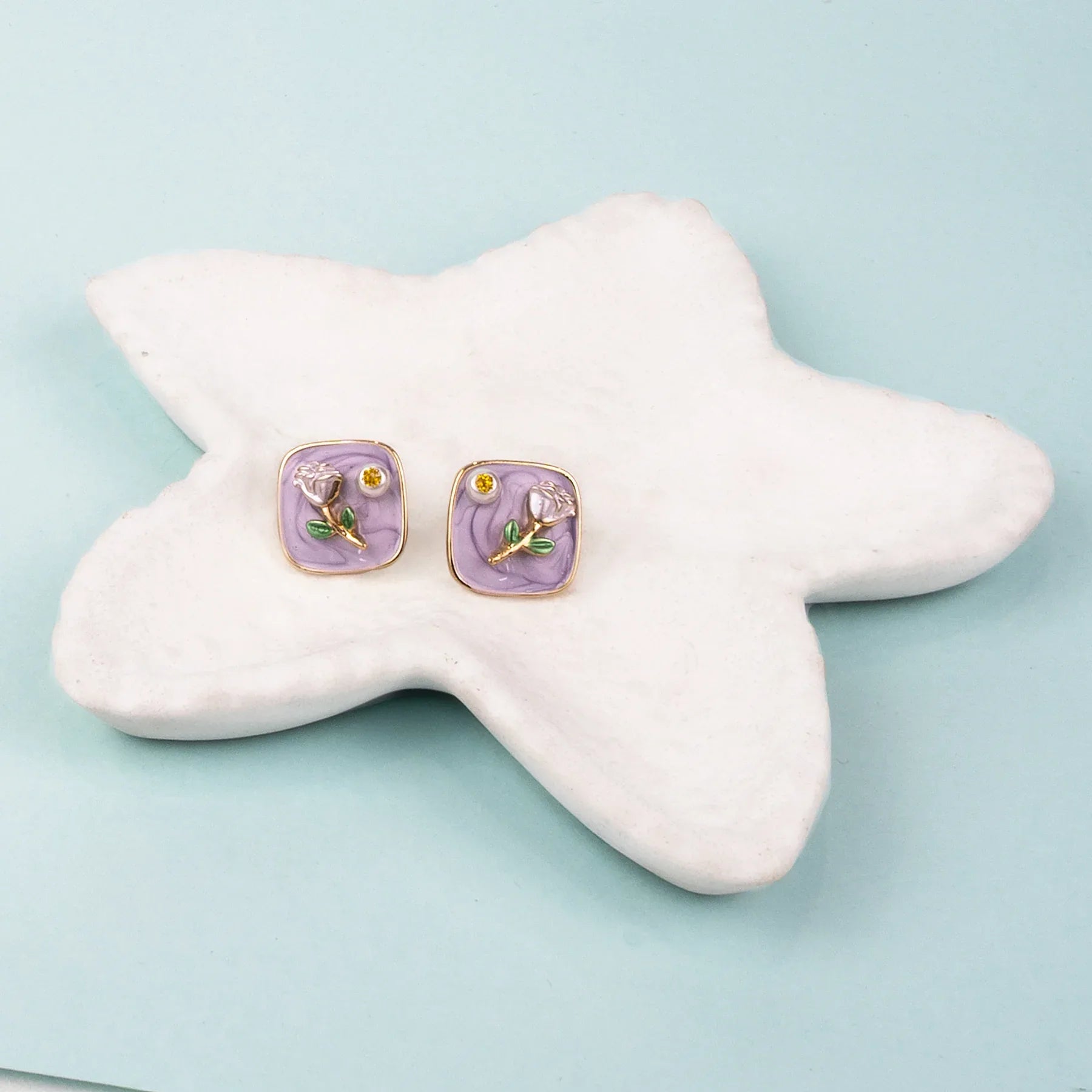 Pair of square earrings with floral design on a star-shaped white stand against a light blue background