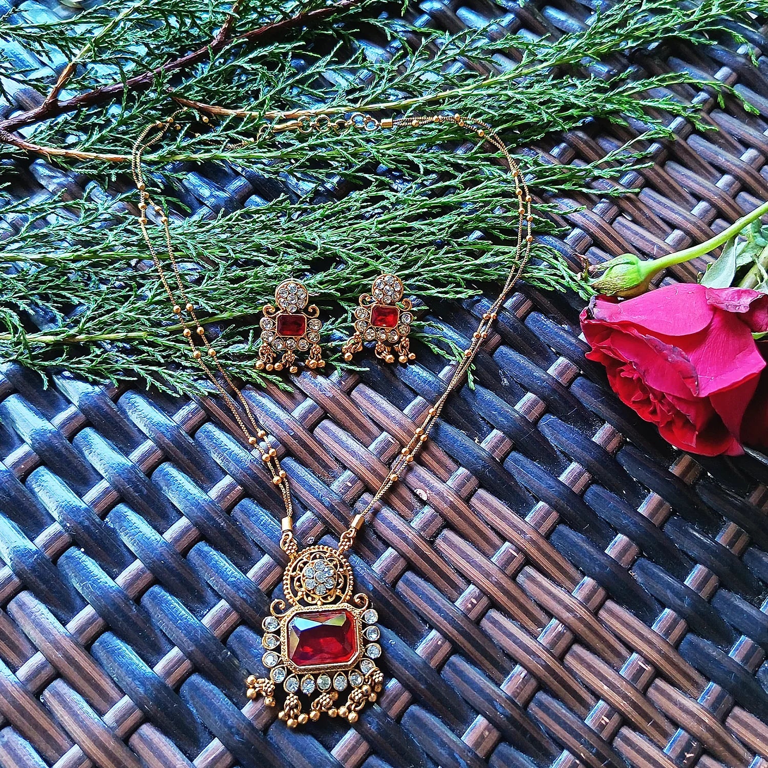 Gold necklace with red gemstone and matching earrings on a textured surface with greenery and a pink rose.