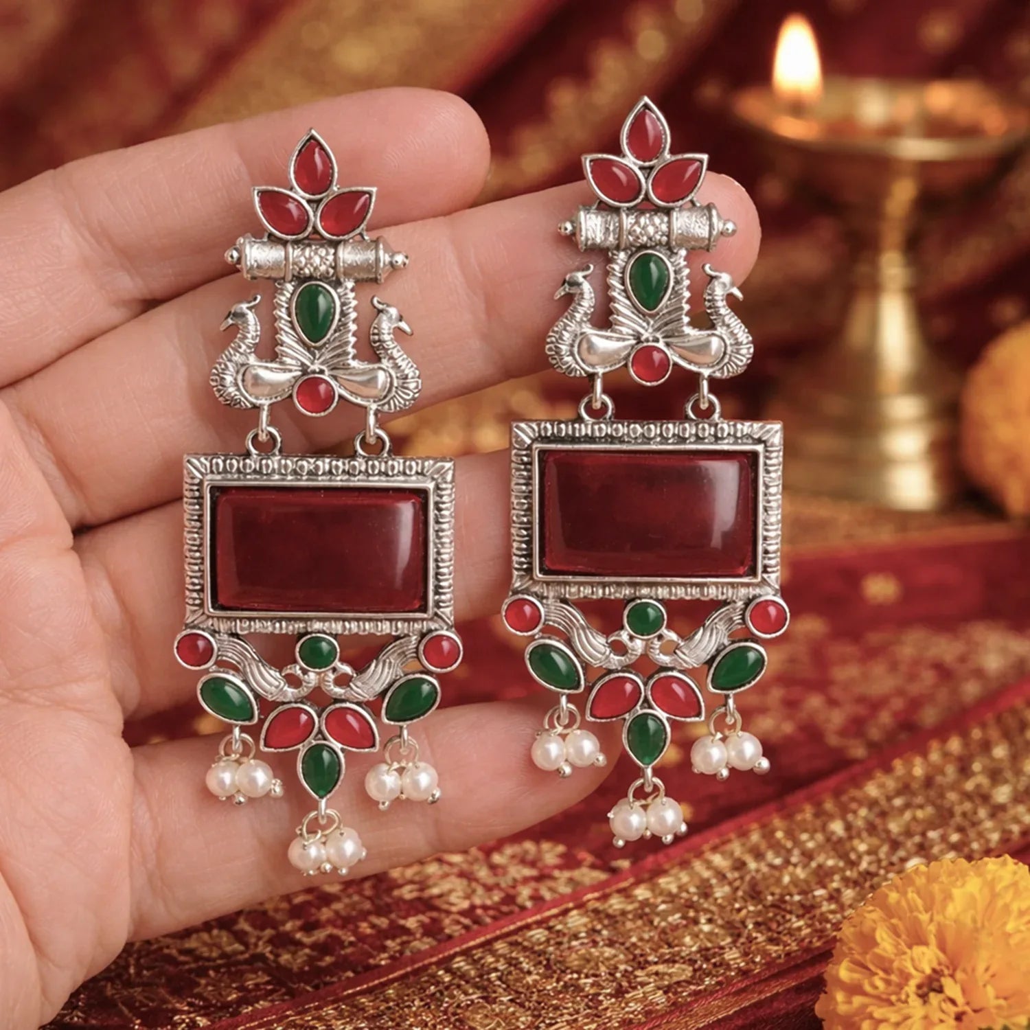 Ornate earrings with red, green, and silver design held by a hand on a decorative background.