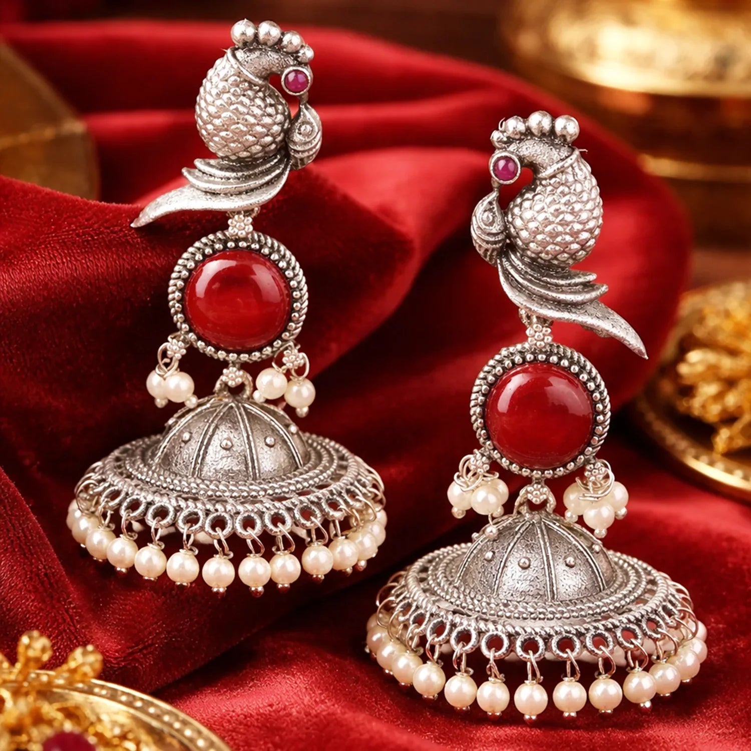 Ornate silver earrings with red stones and pearl-like beads on a red fabric background