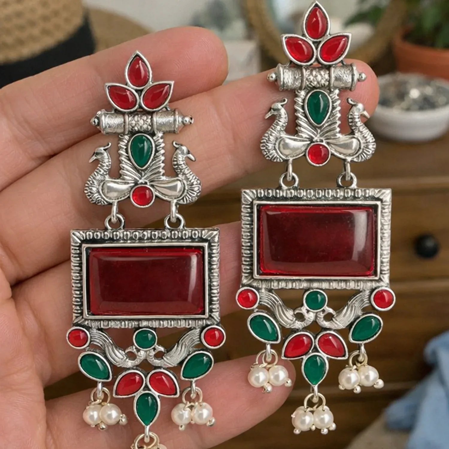 Ornate earrings with red and green stones held in a hand against a blurred background