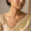 Woman wearing a gold necklace with a pendant, dressed in a beige saree.
