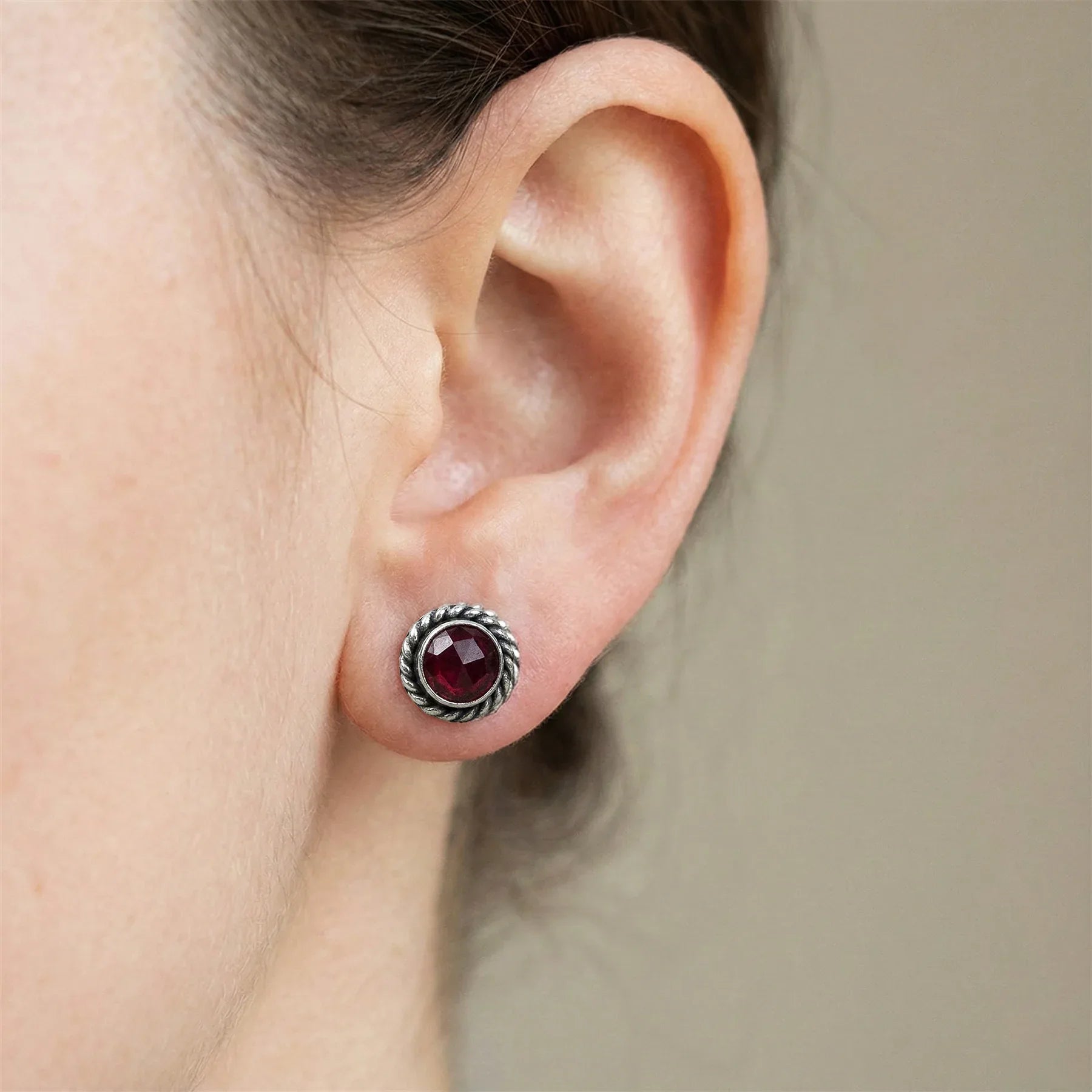 Close-up of an ear wearing a silver earring with a red gemstone on a neutral background