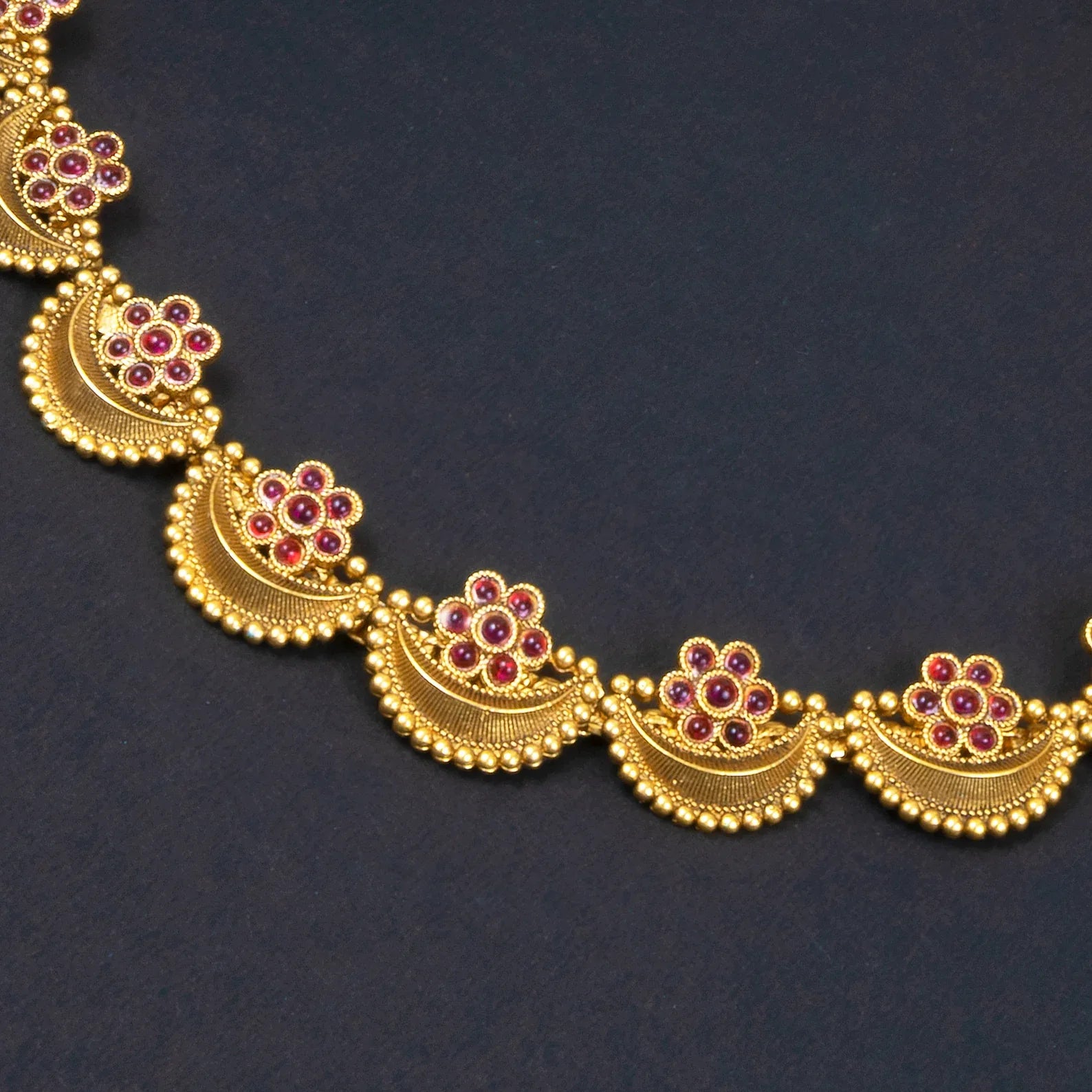 Gold necklace with floral designs and red gemstones on a dark background