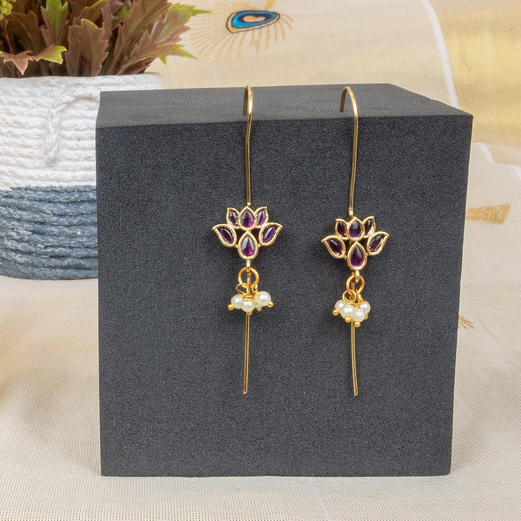 Gold lotus-shaped earrings with purple gemstones and pearl accents on a black stand.