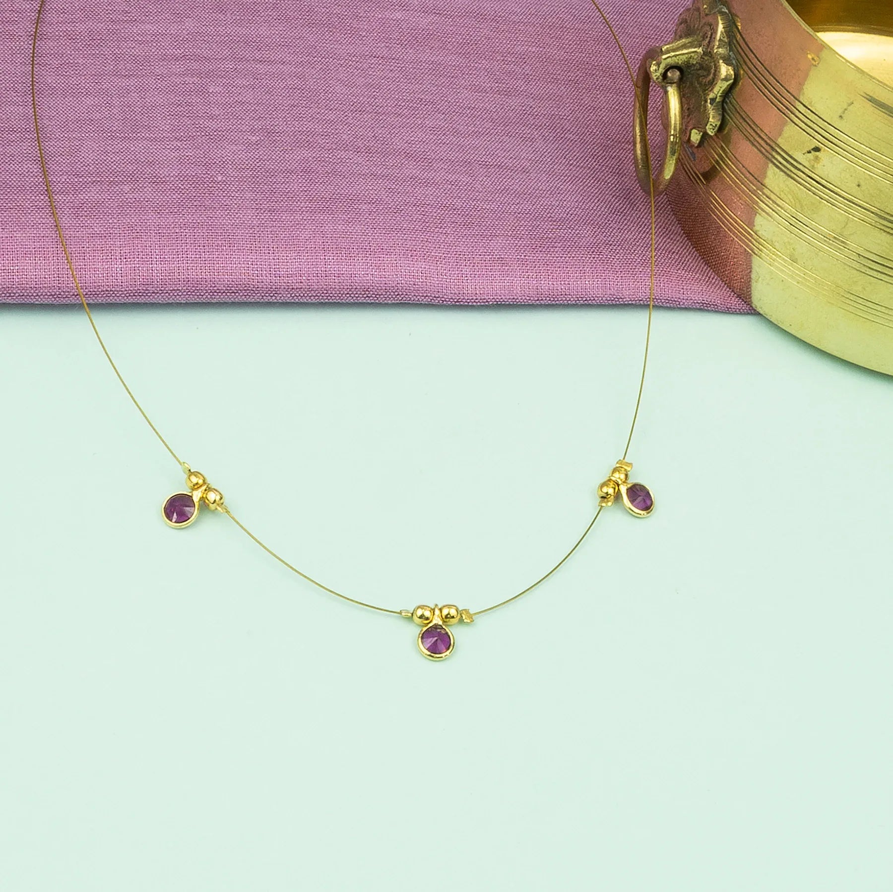 Gold necklace with purple gemstones on a light green surface with a pink fabric background