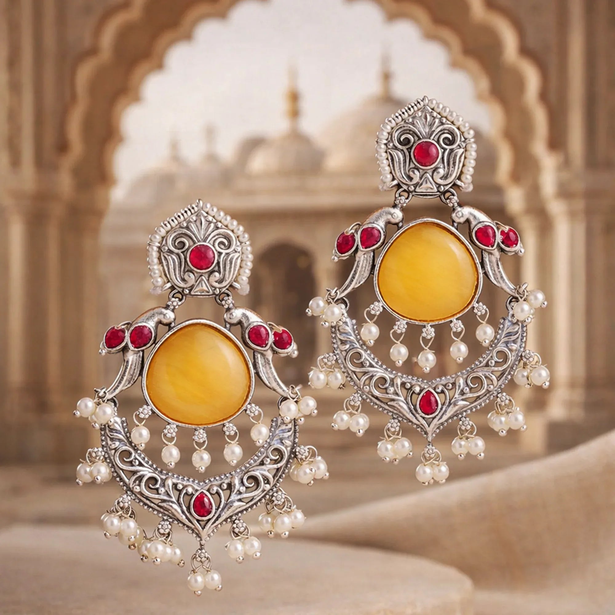 Ornate earrings with yellow centers, red stones, and pearl accents against a blurred architectural background.